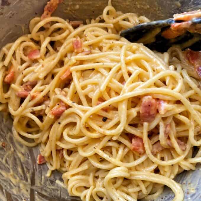 Spaghetti Carbonara A close-up of spaghetti mixed with a creamy sauce and pieces of cooked bacon or ham, being stirred with a black utensil.