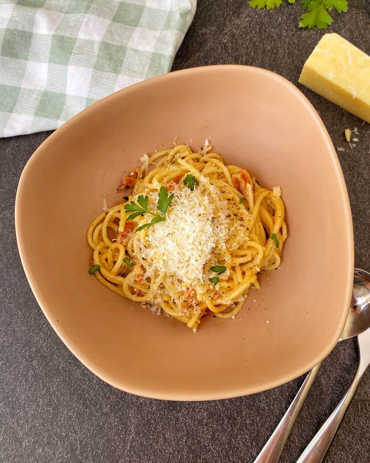 A bowl of spaghetti topped with grated cheese and parsley, placed on a dark surface next to a block of cheese, a checkered cloth, and two spoons.