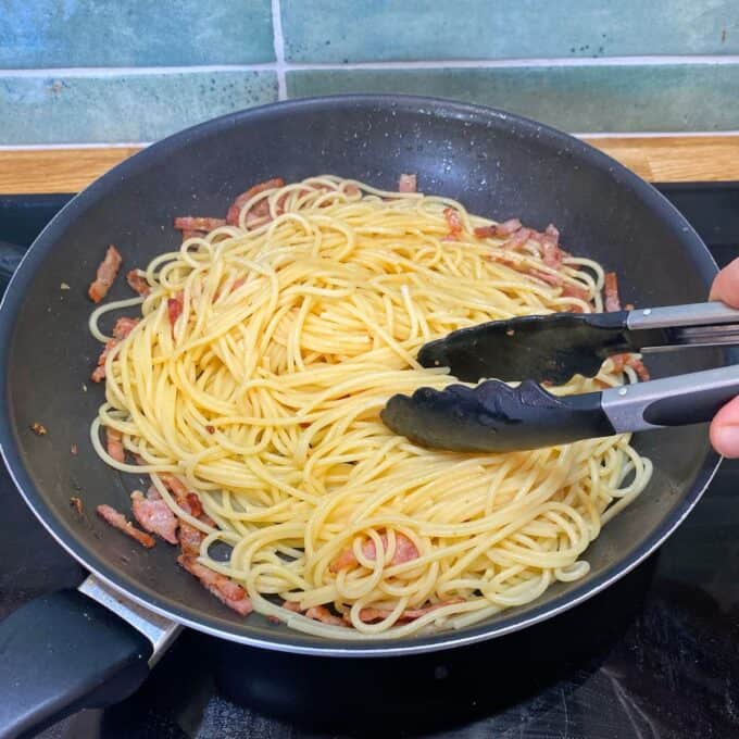 Spaghetti Carbonara A pan with cooked spaghetti and pieces of bacon is being stirred with metal tongs on a stovetop.