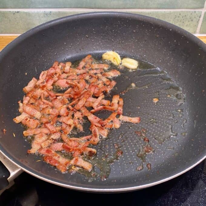 Spaghetti Carbonara Bacon strips cooking in a frying pan with two cloves of garlic, some oil, and visible rendered fat.