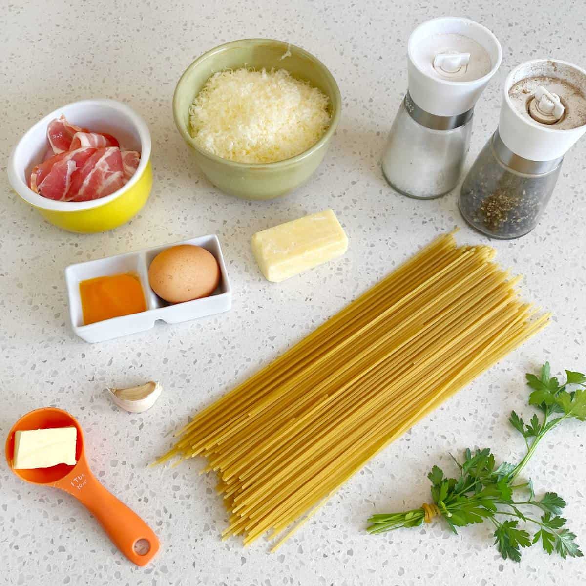 Ingredients for pasta Carbonara arranged on a counter: spaghetti, egg, cheese, pancetta, butter, garlic, parsley, salt, and pepper.