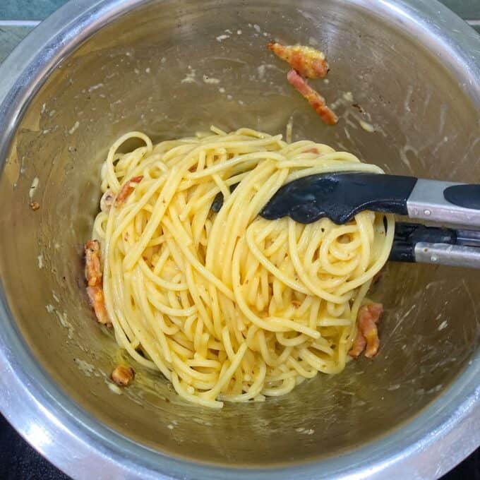 Spaghetti Carbonara A stainless steel bowl containing spaghetti with pieces of bacon or pancetta, coated in a creamy sauce, with a pair of kitchen tongs resting inside.