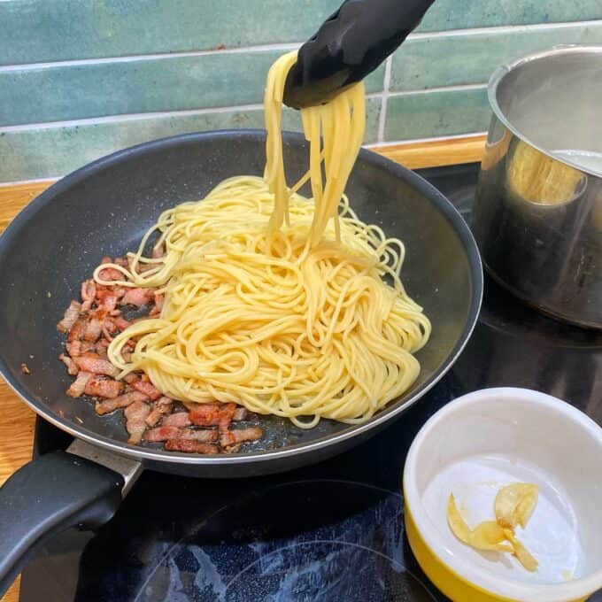Spaghetti Carbonara Cooked spaghetti is being added to a frying pan with bacon using tongs. A pot of water and a bowl with garlic cloves are nearby on the stovetop.