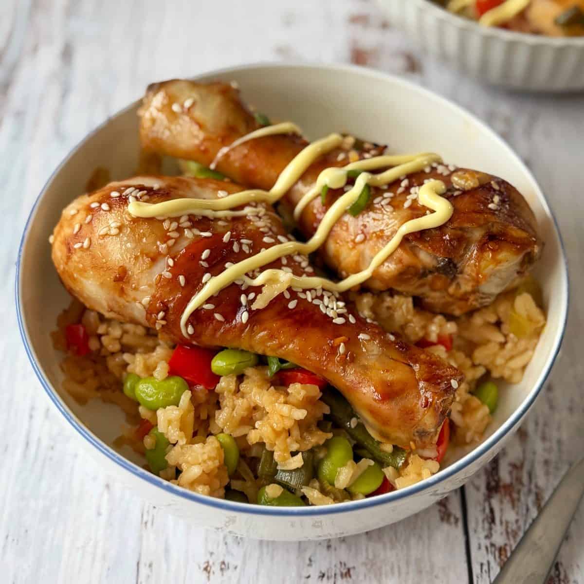 Teriyaki Chicken Drumsticks A bowl of rice with mixed vegetables, topped with two glazed chicken drumsticks, drizzled with sauce, and sprinkled with sesame seeds.