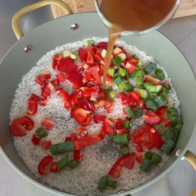 Teriyaki Chicken Drumsticks Chopped red bell peppers and green onions on uncooked rice in a pot as broth is being poured in.