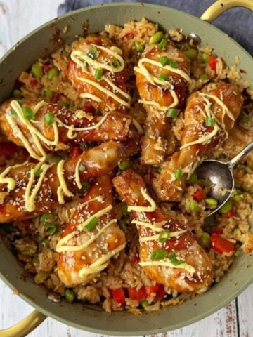 A pan filled with rice, vegetables, and glazed chicken drumsticks, topped with a drizzle of sauce, sesame seeds, and chopped green onions, with a spoon on the side.
