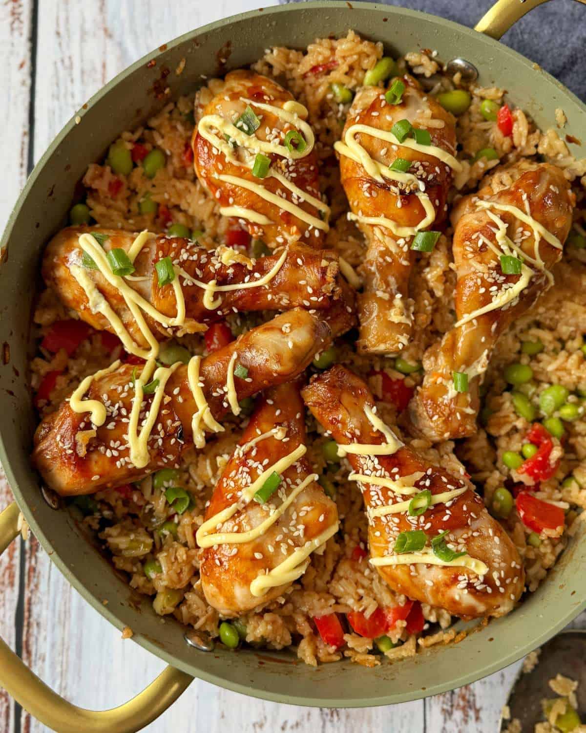 Teriyaki Chicken Drumsticks A pan filled with seasoned chicken drumsticks on top of fried rice with vegetables, garnished with sesame seeds, chopped green onions, and a drizzle of sauce.
