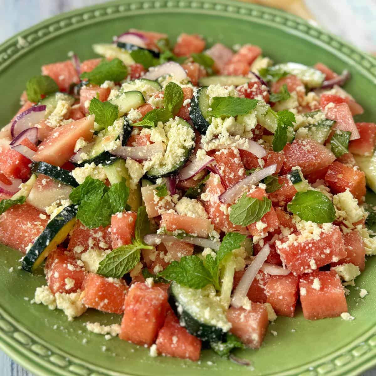 Watermelon, Feta and Mint Salad A green plate with a salad of watermelon cubes, cucumber slices, red onion, mint leaves, and crumbled cheese.