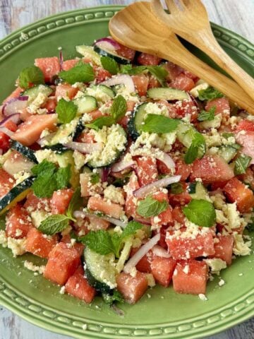 A green plate with a salad of watermelon cubes, cucumber slices, red onion, crumbled cheese, and mint leaves, with wooden salad servers on the side.