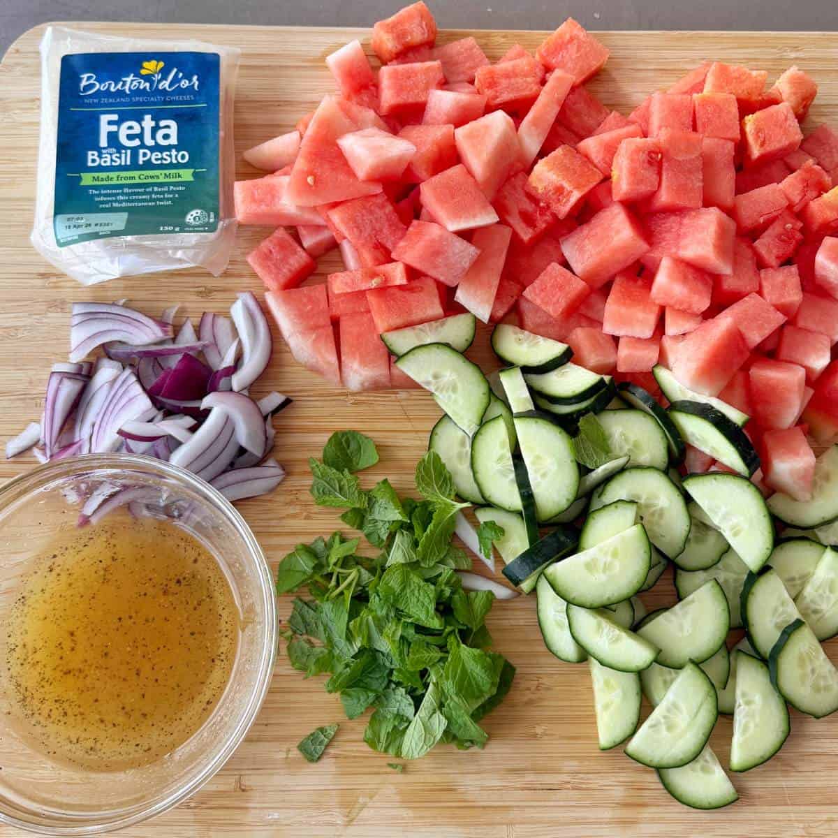 Watermelon, Feta and Mint Salad Chopped watermelon, cucumber, red onion, mint, basil pesto feta, and a bowl of vinaigrette are arranged on a wooden cutting board.