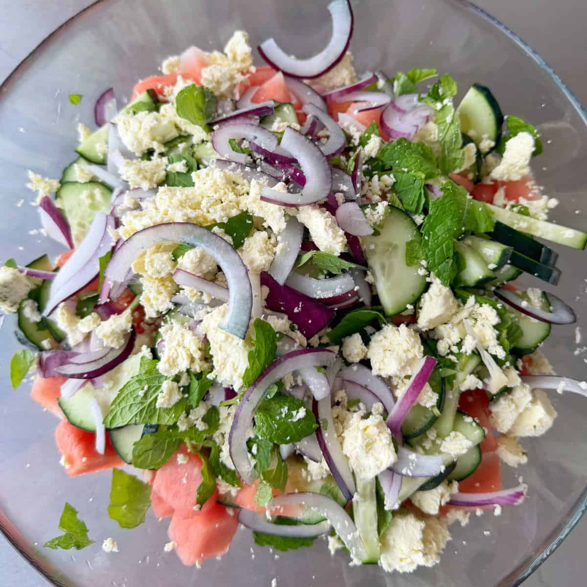 Watermelon, Feta and Mint Salad A bowl of salad containing sliced cucumbers, red onions, watermelon, crumbled feta cheese, and fresh mint leaves.