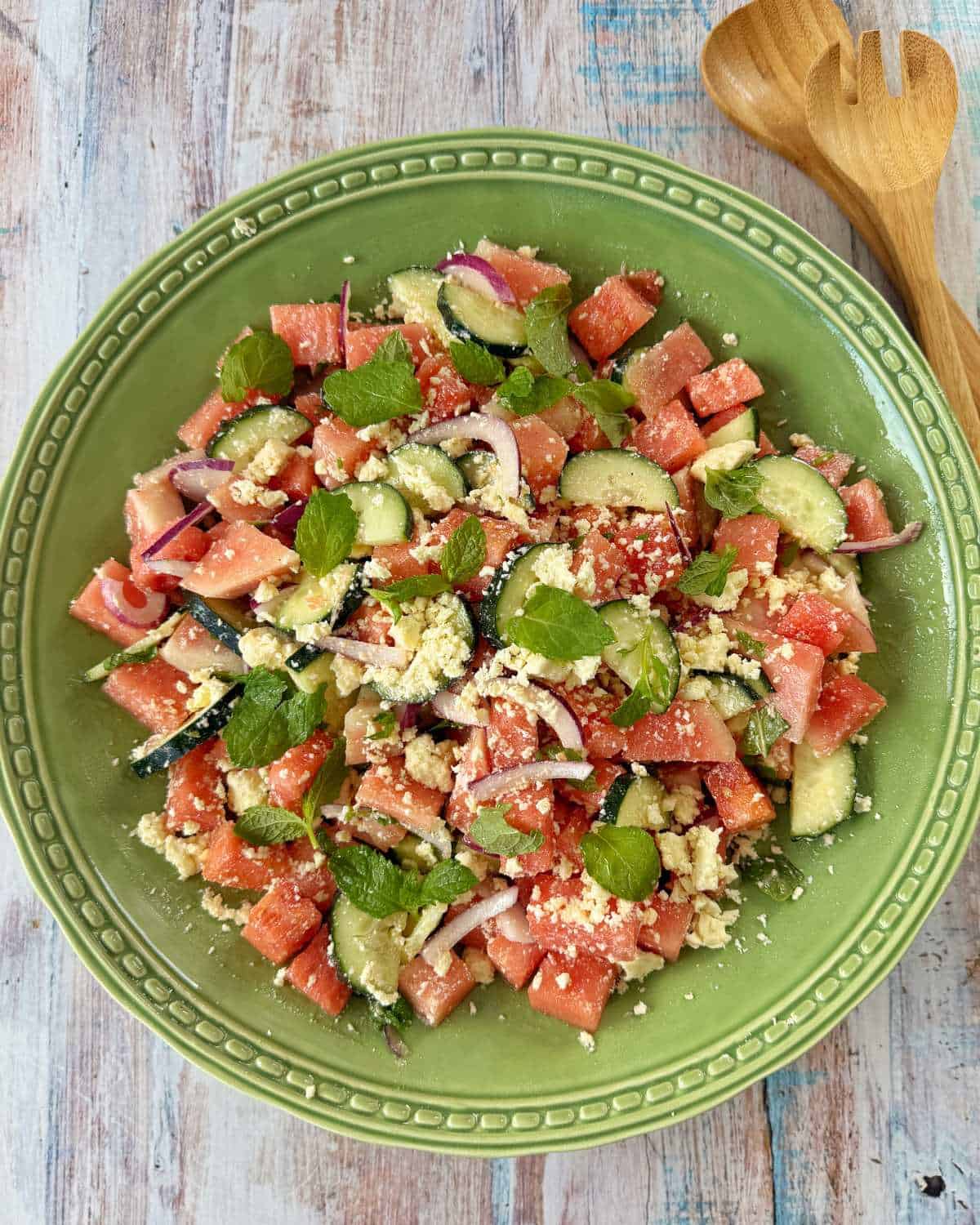 Watermelon, Feta and Mint Salad A green plate filled with watermelon salad containing cucumber, red onion, feta cheese, and mint leaves; wooden salad utensils are nearby.