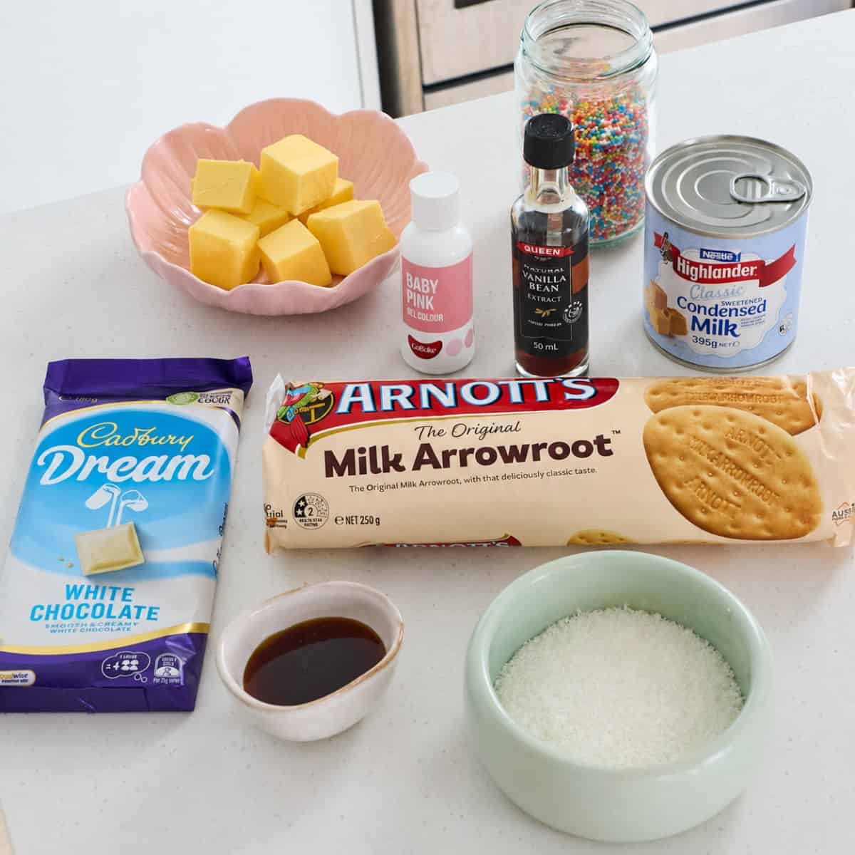 Sprinkle Biscuit Slice A variety of baking ingredients on a counter, perfect for making Sprinkle Biscuit Slice, including butter, sprinkles, condensed milk, vanilla, white chocolate, biscuits, food coloring, and desiccated coconut.