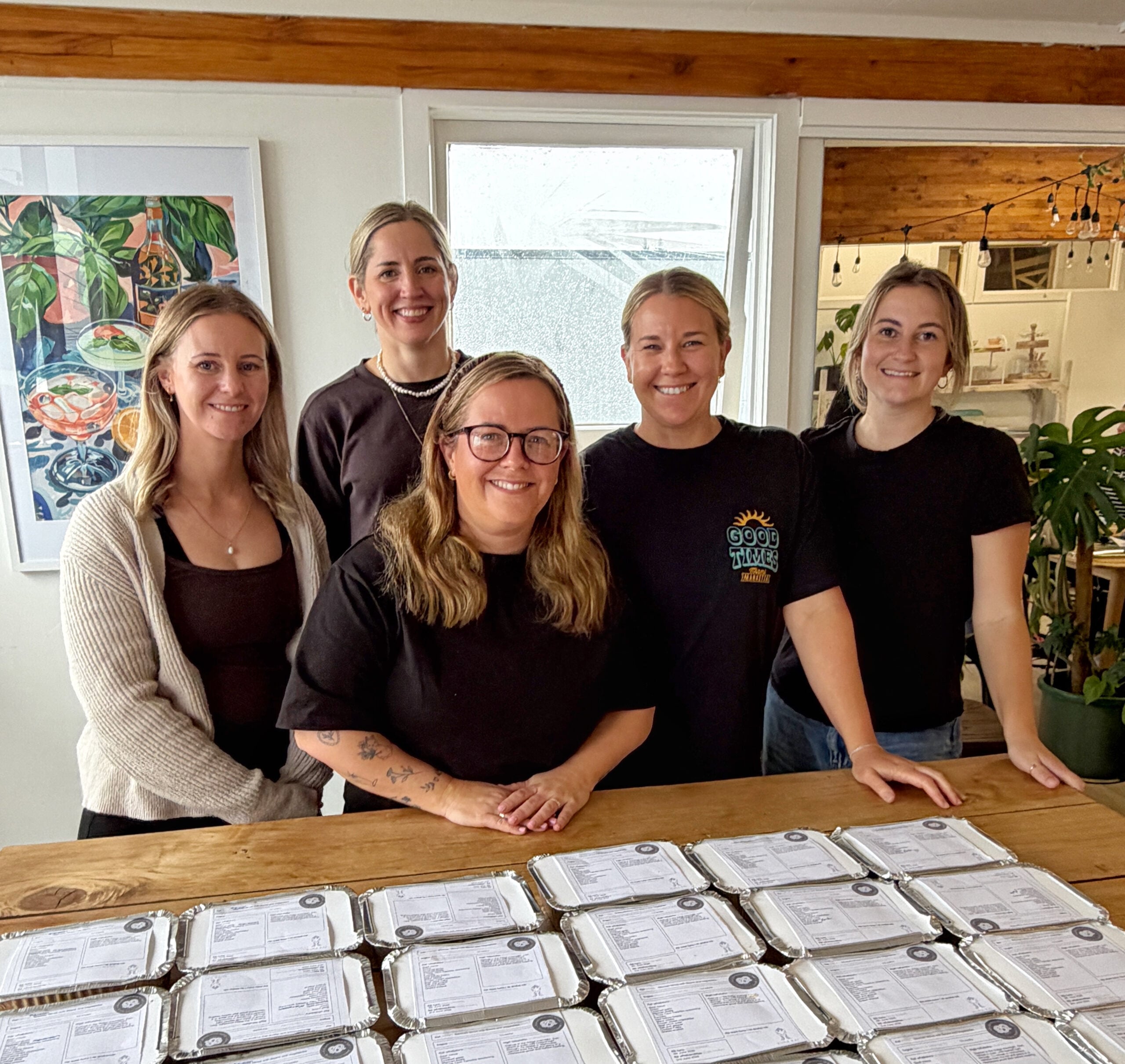 Five women stand behind a table filled with prepared meal containers, smiling at the camera in a well-lit room with artwork and plants.