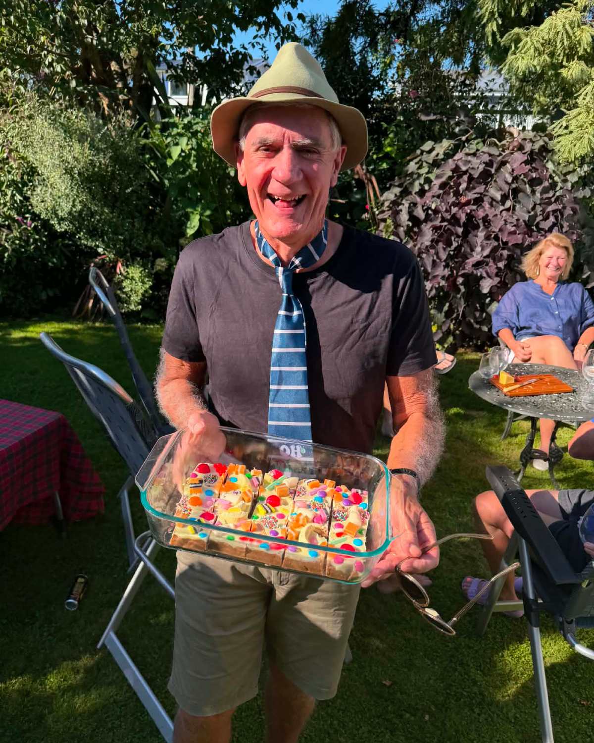 XL Pick and Mix Lolly Slice A man wearing a hat and tie stands outdoors, smiling and holding a glass dish with a decorated cake; people and garden furniture are visible in the background.