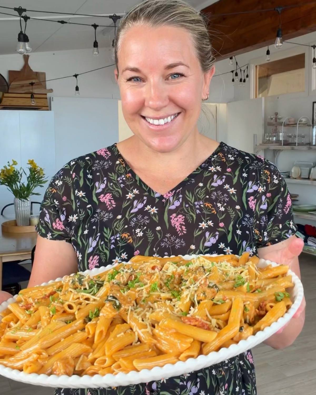 A woman in a floral dress holds a large oval platter of Marry Me Chicken Pasta topped with sauce, herbs, and grated cheese in a bright kitchen.
