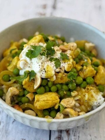 A bowl of One Pot Chicken and Pea Curry with tender chicken pieces, green peas, cashews, a dollop of yogurt, and fresh herbs sits on a white wooden surface.