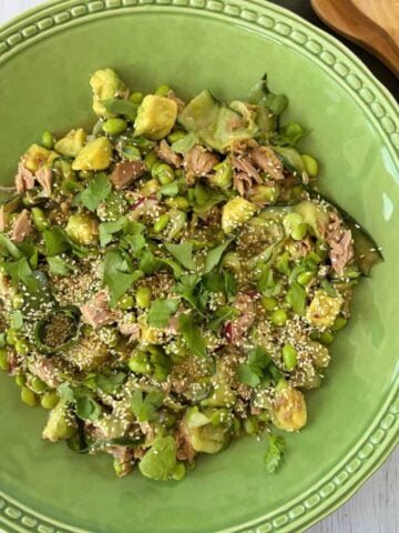 A green bowl filled with a salad of tuna, edamame, gnocchi, fresh herbs, and sesame seeds, with wooden serving utensils beside it.