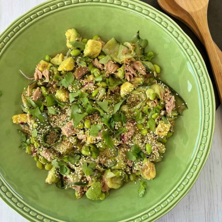 A green bowl filled with a salad of tuna, edamame, gnocchi, fresh herbs, and sesame seeds, with wooden serving utensils beside it.