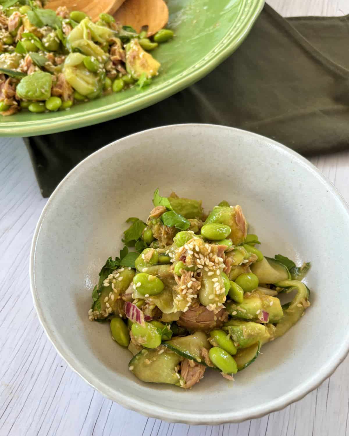 Zesty Tuna, Avocado and Edamame Salad A bowl of salad with cucumbers, edamame, red onion, greens, tuna, and sesame seeds, with a larger serving bowl and wooden utensils in the background.
