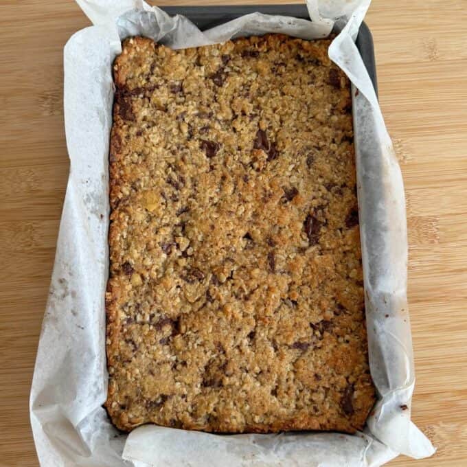Banana and Dark Chocolate Bumper Bars A rectangular baking tray lined with parchment paper holds a baked oat and chocolate chip bar on a wooden surface.