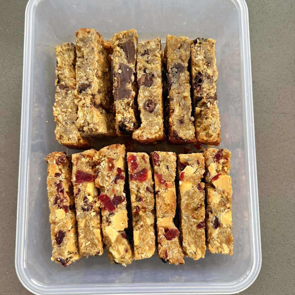 Banana and Dark Chocolate Bumper Bars A plastic container holds two rows of homemade oat bars, some with chocolate chunks and others with visible dried fruit pieces.