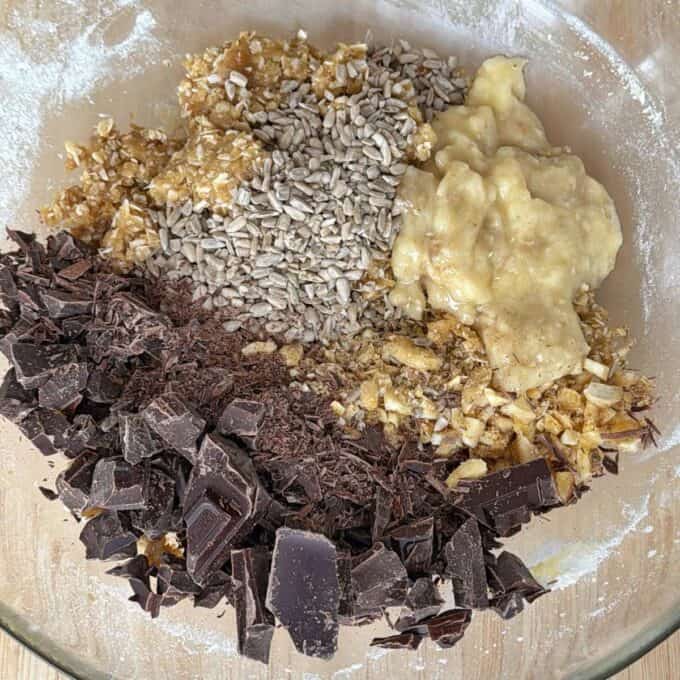 Banana and Dark Chocolate Bumper Bars A glass bowl containing chopped dark chocolate, sunflower seeds, mashed banana, oats, and what appears to be chopped nuts and honey.