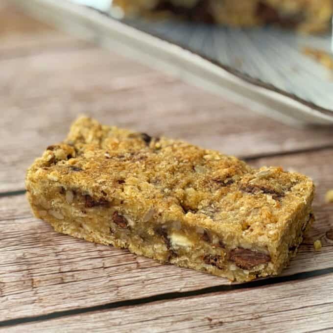 Banana and Dark Chocolate Bumper Bars A close-up of a homemade granola bar with visible oats, nuts, and chocolate chunks on a wooden surface; a plate with more bars is in the background.
