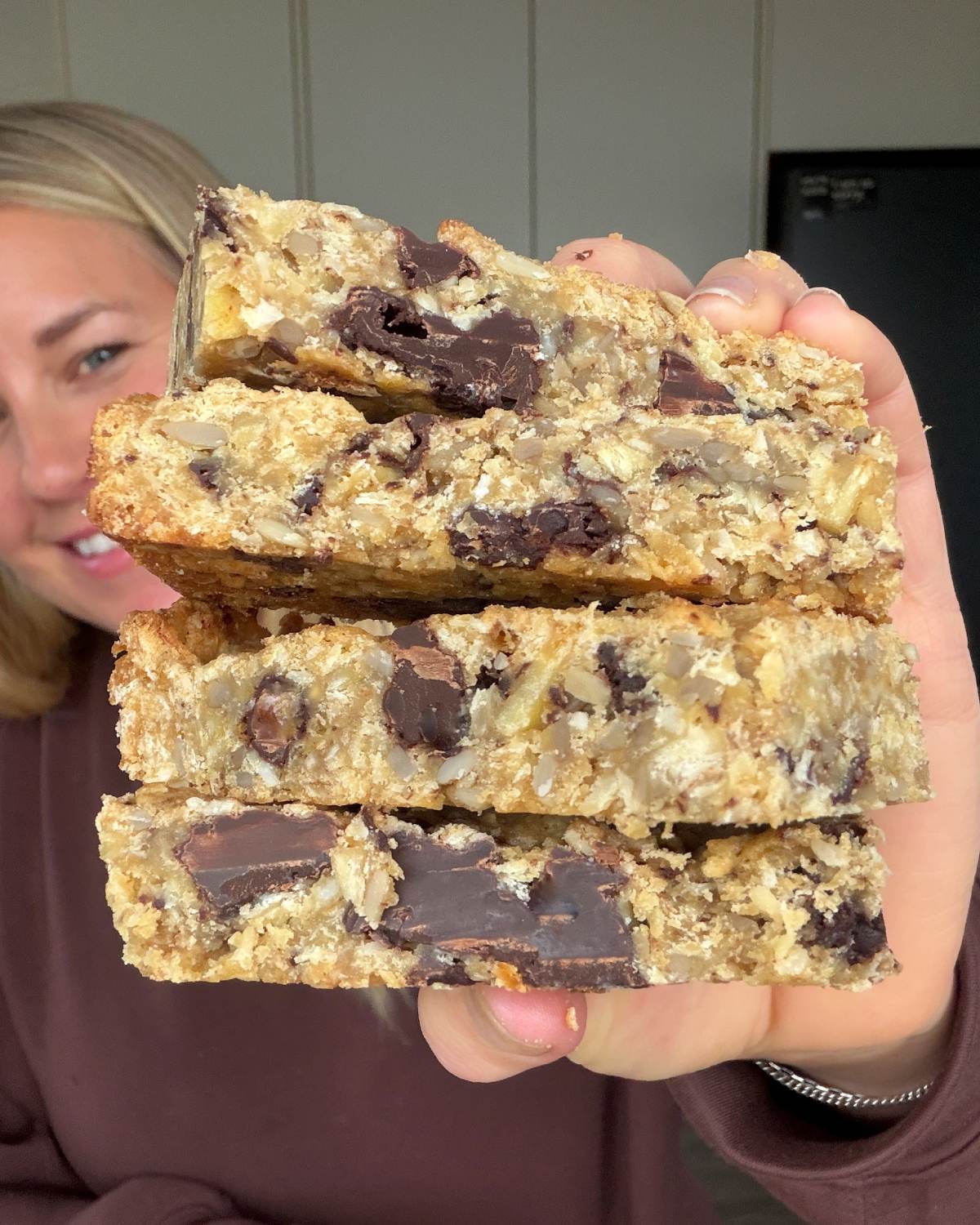 Banana and Dark Chocolate Bumper Bars A person holds up a stack of four thick, homemade oat bars with visible chunks of chocolate.