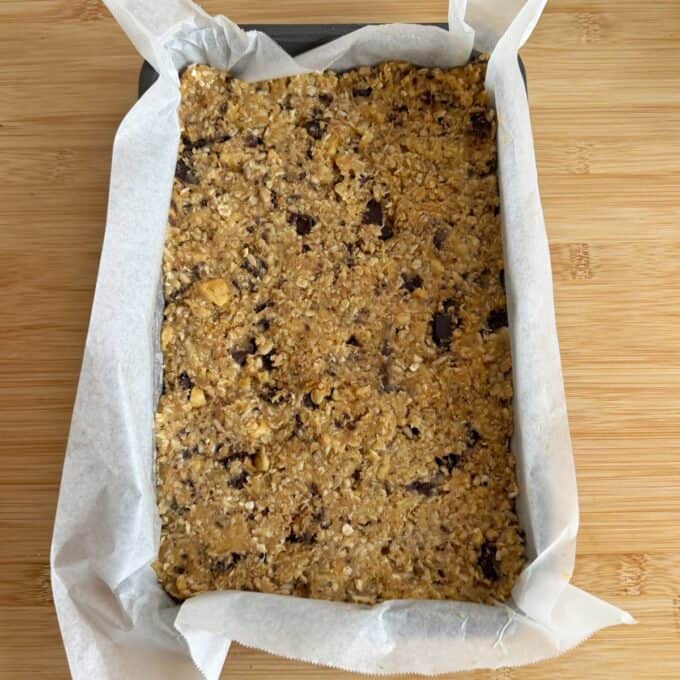 Banana and Dark Chocolate Bumper Bars A parchment-lined baking pan filled with unbaked oat and chocolate chip cookie dough sits on a wooden surface.