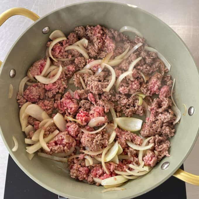 Beef Mince Chow Mein Ground beef and sliced onions cooking in a large pan on a stovetop, with some meat still pink and some browned.