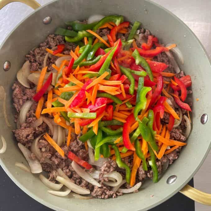 Beef Mince Chow Mein Sliced onions, ground beef, and julienned carrots, red bell peppers, and green bell peppers cooking in a large pan.