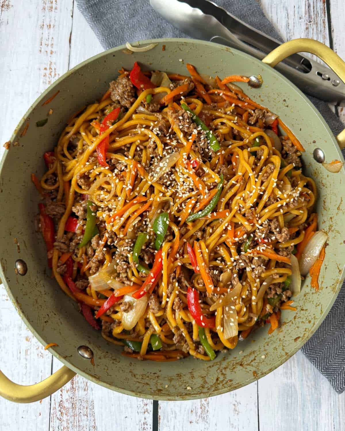 Beef Mince Chow Mein A large pan filled with stir-fried noodles, ground beef, bell peppers, onions, carrots, and sesame seeds. Metal tongs rest nearby on a gray cloth.