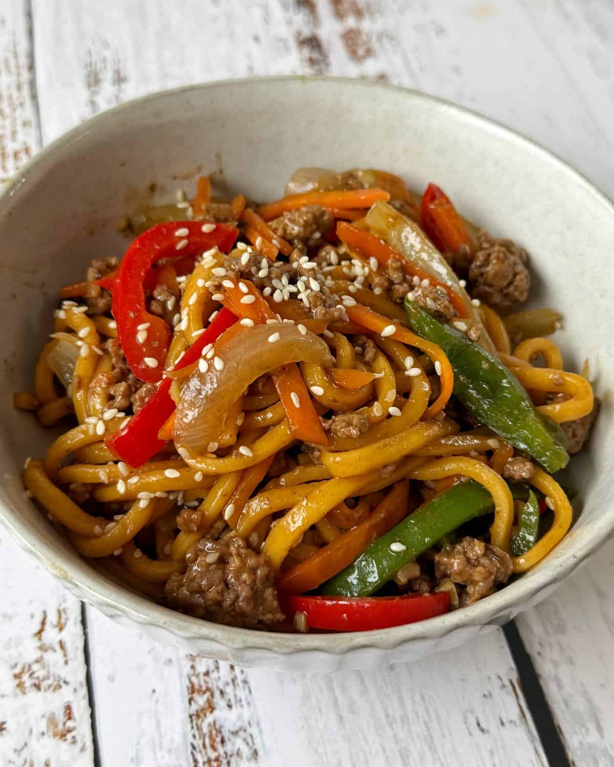 Beef Mince Chow Mein A bowl of stir-fried noodles with ground meat, red and green bell peppers, onions, carrots, and sesame seeds on top.