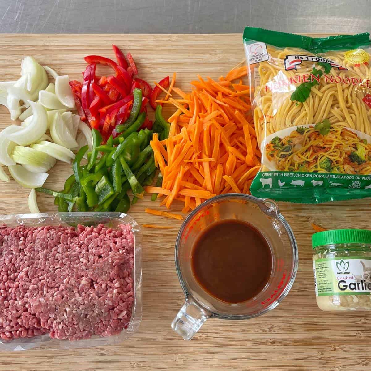 Beef Mince Chow Mein Chopped onions, red and green bell peppers, and carrots on a cutting board with ground beef, noodles, sauce in a measuring cup, and a garlic jar.