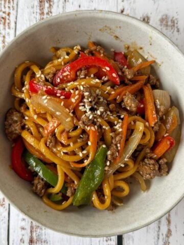A bowl of stir-fried noodles with ground meat, sliced red peppers, onions, green peppers, carrots, and sesame seeds on top, placed on a rustic white wooden surface.