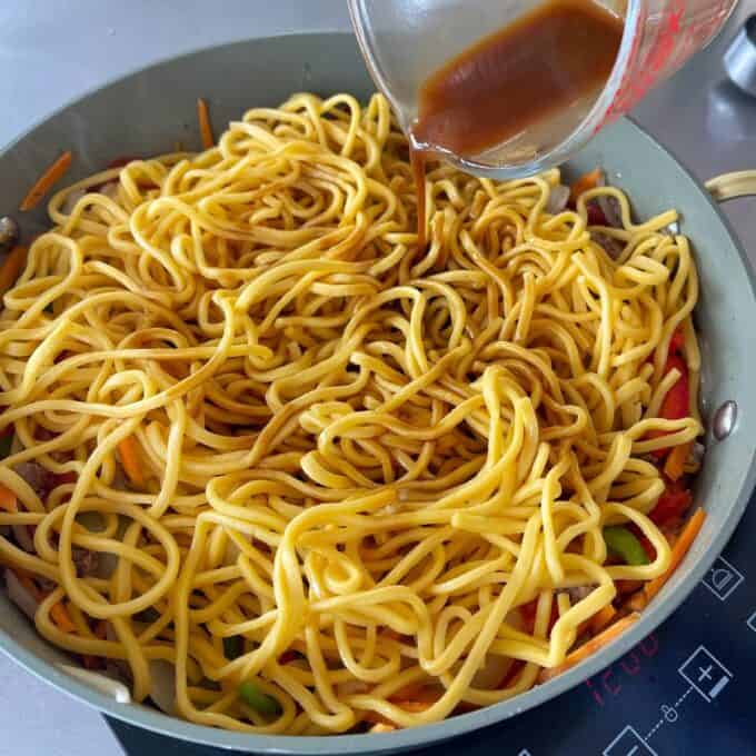 Beef Mince Chow Mein A measuring cup pours sauce over cooked noodles with vegetables in a frying pan on an induction stove.