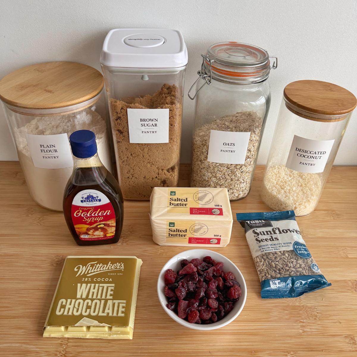 Berry and White Chocolate Bumper Bars Ingredients displayed on a wooden surface: plain flour, brown sugar, oats, desiccated coconut, golden syrup, salted butter, white chocolate, dried cranberries, and sunflower seeds.