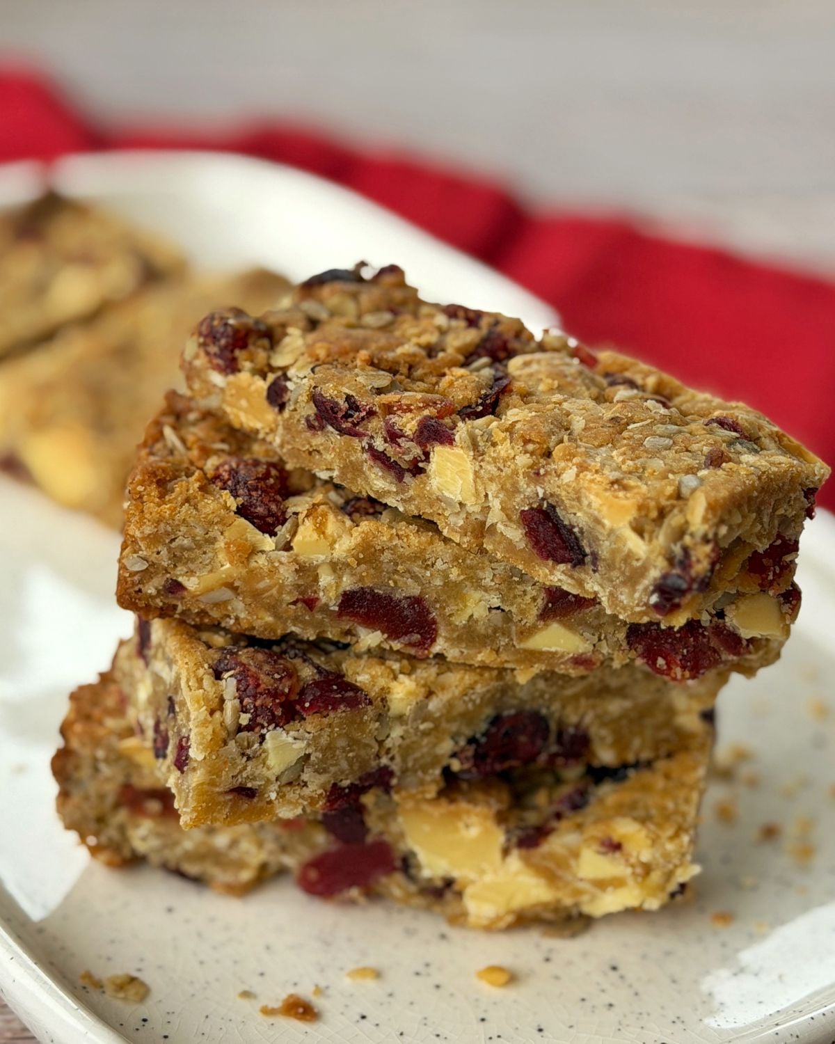 Berry and White Chocolate Bumper Bars Three stacked oat bars with visible dried cranberries and nuts on a white plate, with more bars and a red cloth in the background.