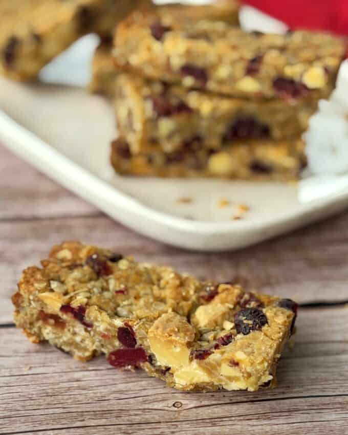 Berry and White Chocolate Bumper Bars Close-up of a homemade granola bar with visible dried fruit and nuts on a wooden surface, with more bars stacked on a plate in the background.