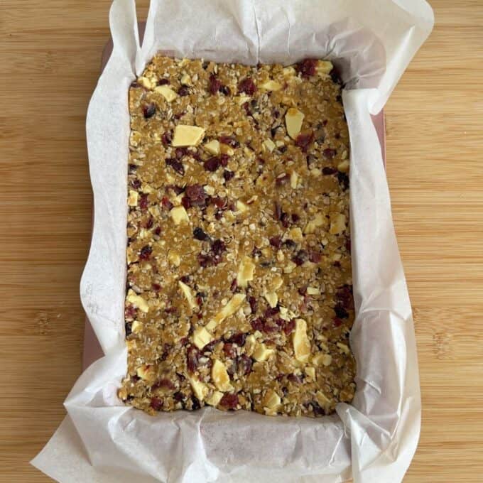 Berry and White Chocolate Bumper Bars A rectangular pan lined with parchment paper filled with an unbaked oat, nut, and dried fruit mixture, ready to be baked.