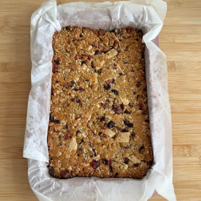 Berry and White Chocolate Bumper Bars A rectangular oat and fruit flapjack slab sits in a baking tray lined with parchment paper on a wooden surface.