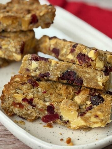 Five oat and nut granola bars with visible dried berries stacked on a white speckled plate, with a red cloth in the background.