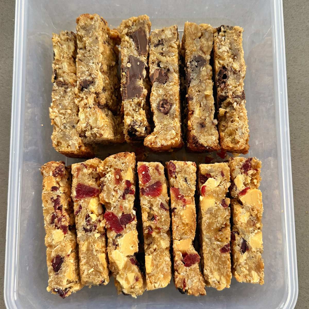Twelve homemade granola bars with visible nuts, dried fruit, and chocolate pieces arranged in two rows inside a clear plastic container.