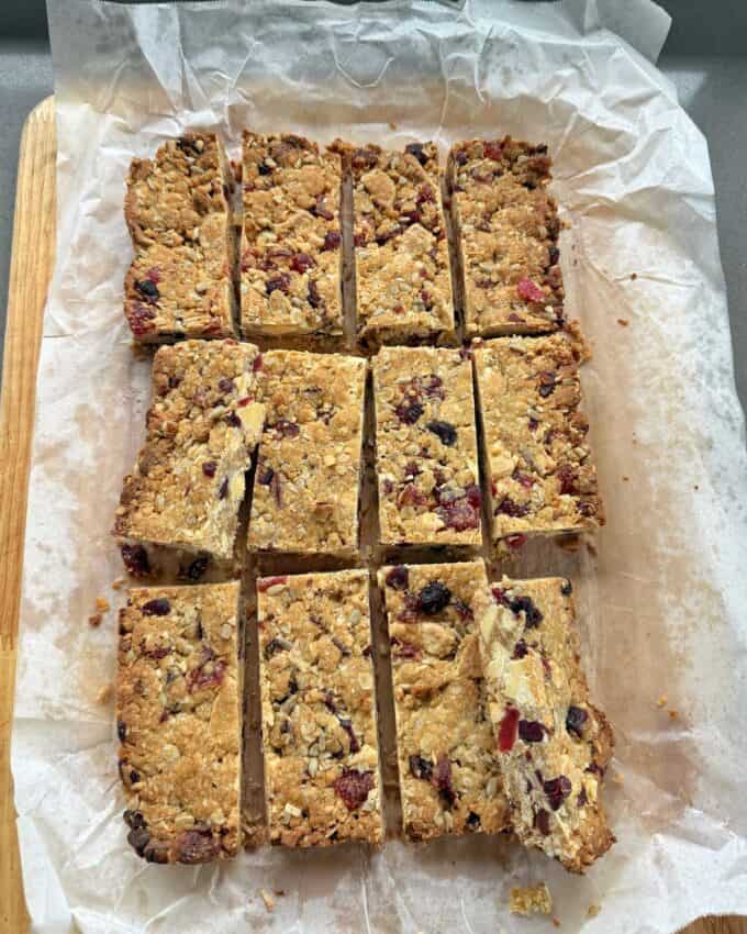 Berry and White Chocolate Bumper Bars A tray of homemade oat and fruit flapjacks cut into twelve rectangular pieces, with one piece slightly separated from the rest, on parchment paper.