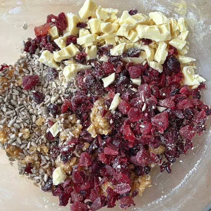 Berry and White Chocolate Bumper Bars Chopped white chocolate, dried cranberries, sunflower seeds, and raisins arranged in a mixing bowl, partially surrounded by flour.