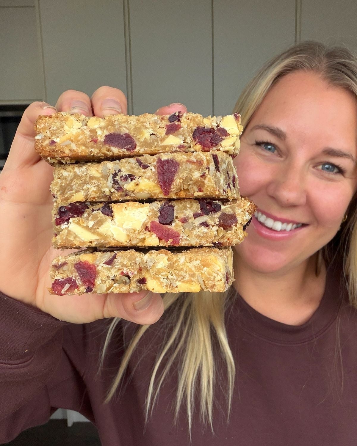 Berry and White Chocolate Bumper Bars A woman holds up a stack of four homemade granola bars with visible nuts and dried fruit, smiling at the camera.