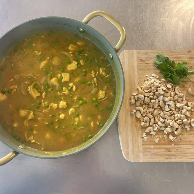 Chicken and Pea Curry A pot of yellow curry with peas and tofu sits beside a cutting board with chopped cashews and fresh parsley on a kitchen counter.