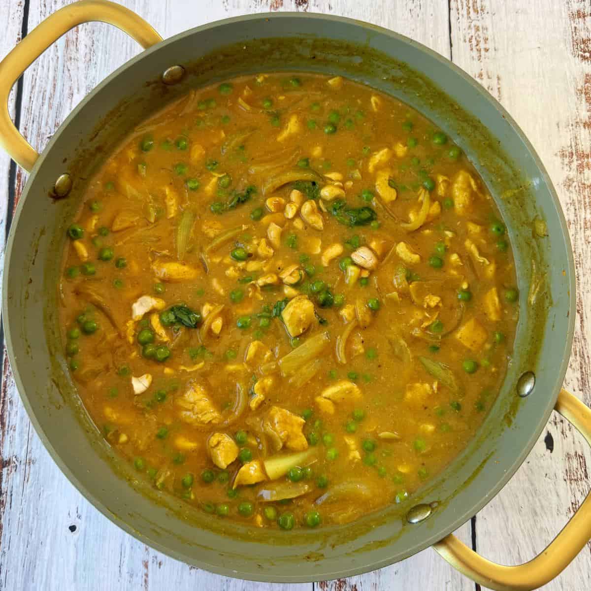 Chicken and Pea Curry Yellow curry with chicken, peas, onions, and spinach simmering in a large green pot with yellow handles on a white wooden surface.