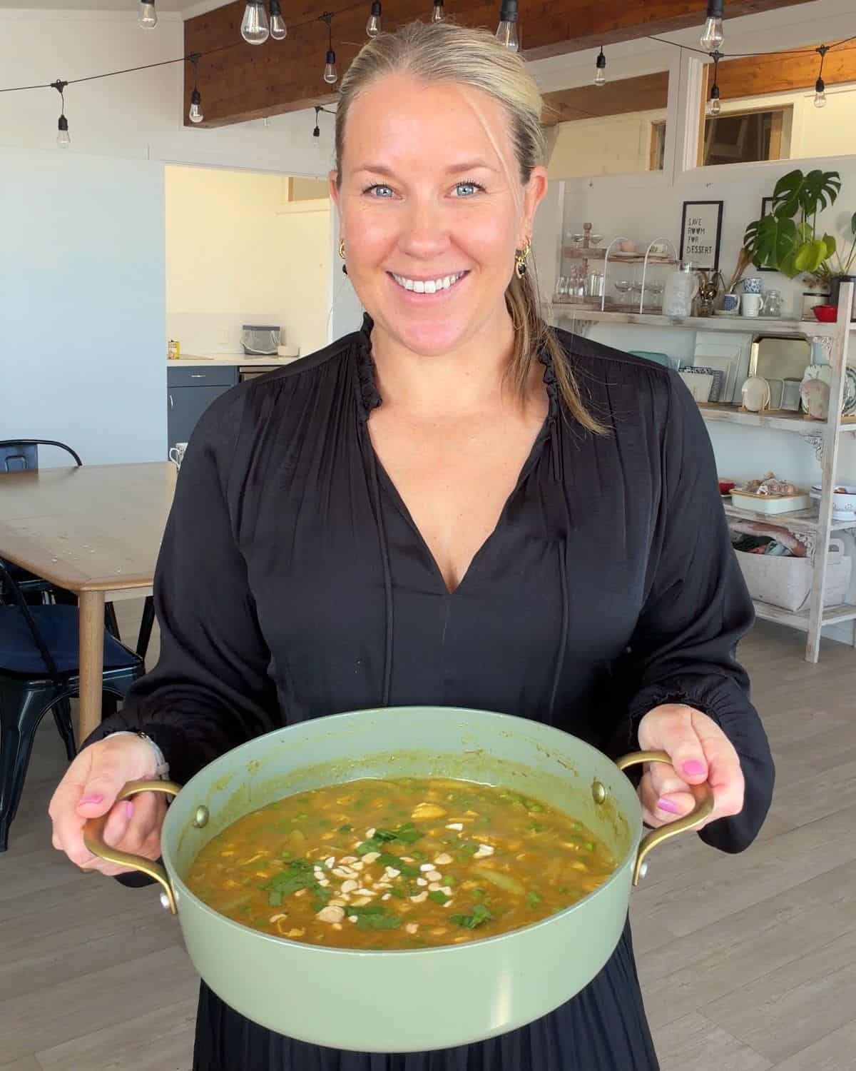 Chicken and Pea Curry A woman in a black dress holds a pot of soup garnished with chopped nuts in a kitchen.